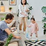 a woman using a mop with her child while her husband is sitting on a couch