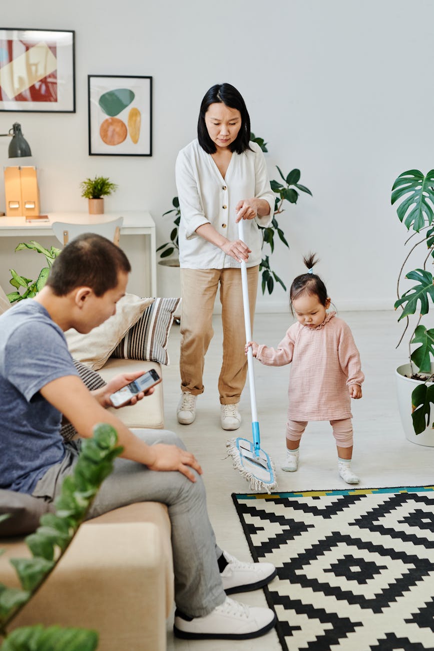 a woman using a mop with her child while her husband is sitting on a couch