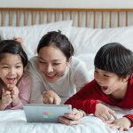 cheerful ethnic mother pointing at tablet screen and children watching and laughing